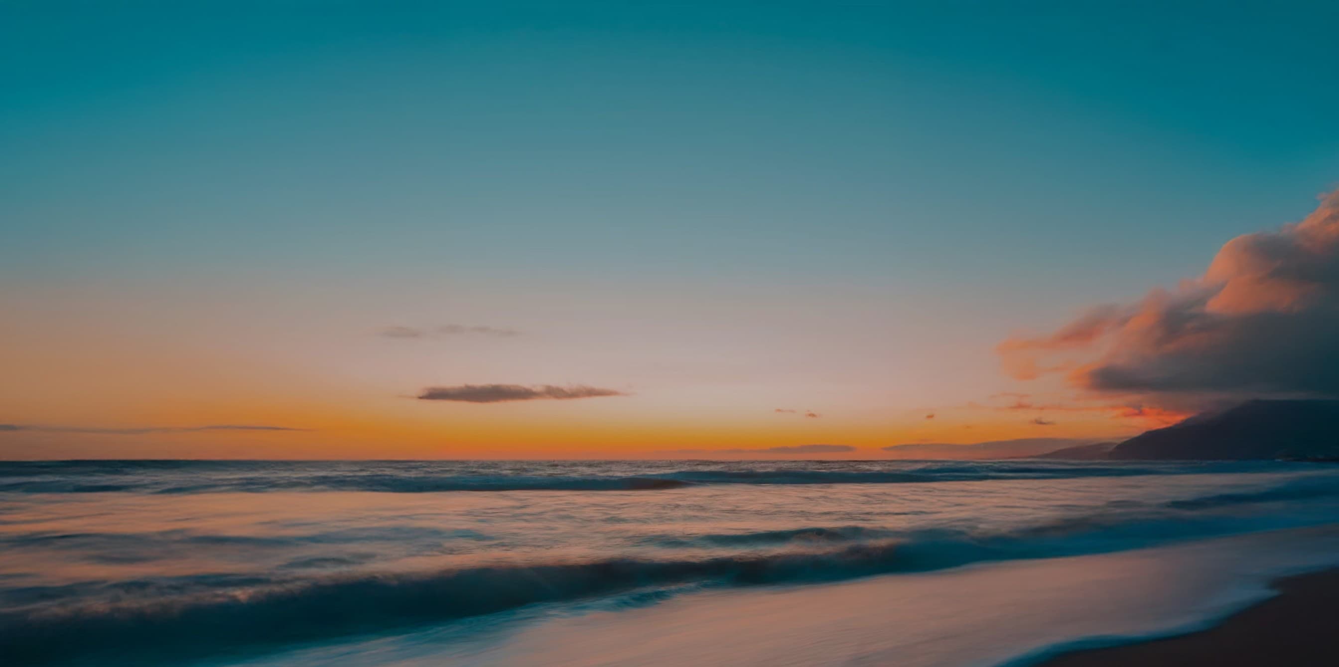 Sandy beach at sunset with gentle waves rolling in, a coastal cliff on the right catching the last warm light of day